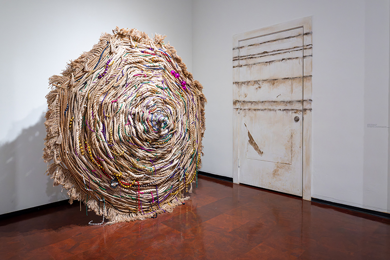 Photograph of gallery with large umbrella created by woven ropes and strings of colorful beads with water lines painted on door