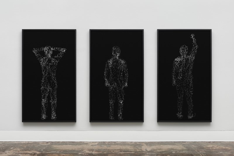 Photograph of three framed artworks installed on white wall. Each piece has a black background and the form a standing figure in white lines