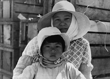 A Japanese mother and child, both wearing head coverings, stand in front of a wooden building.