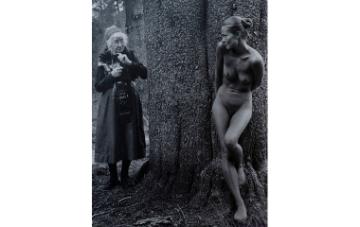 Black and white photograph of old woman carrying a camera and a nude woman standing by a tree looking at each other.