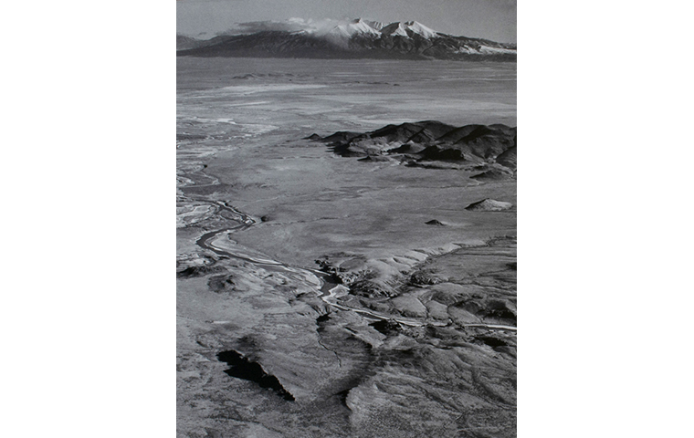 Black and white photograph of aerial view of river valley and mountains. River runs diagonally in the lower part. Snow capped mountains in the distance.