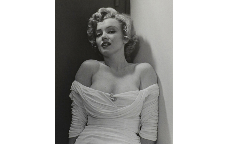 Black and white photograph of Marilyn Monroe looking directly to the viewer. Figure is leaning back and wearing a white dress with a brooch in the center, dress sleeves worn below the shoulder.