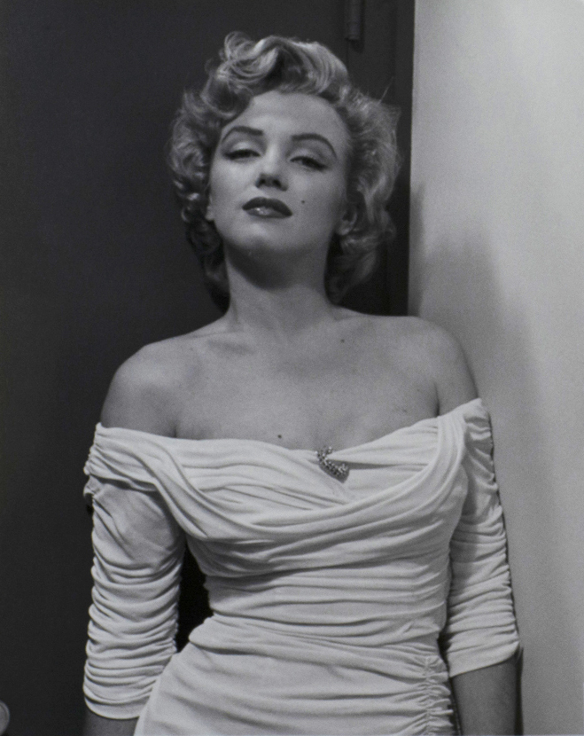 Black and white photograph of Marilyn Monroe looking directly to the viewer. She is leaning to the right and wearing a white dress with a brooch in the center, dress sleeves worn below the shoulder.
