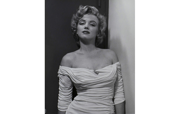 Black and white photograph of Marilyn Monroe looking directly to the viewer. She is leaning to the right and wearing a white dress with a brooch in the center, dress sleeves worn below the shoulder.