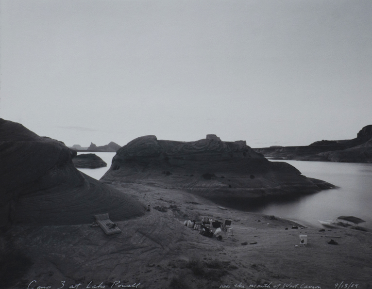 Black and white photograph of treeless and rocky landscape along a lake. Large hill in the center. Folding camping chairs in the center and foreground. An inflatable bed on the left side. A five gallon bucket is on the right. An empty boat docked along the shore on the right side.
