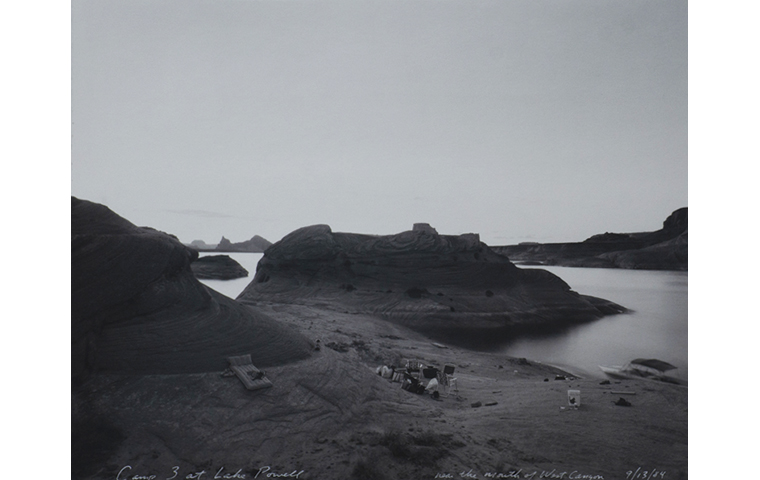 Black and white photograph of treeless and rocky landscape along a lake. Large hill in the center. Folding camping chairs in the center and foreground. An inflatable bed on the left side. A five gallon bucket is on the right. An empty boat docked along the shore on the right side.