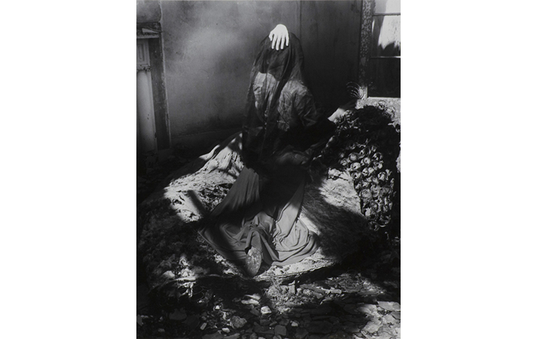 Black and white photograph of figure draped with a sheer veil over their head with their left hand on top. Left side of image is a Greek ionic column. On right side of image is a window frame.