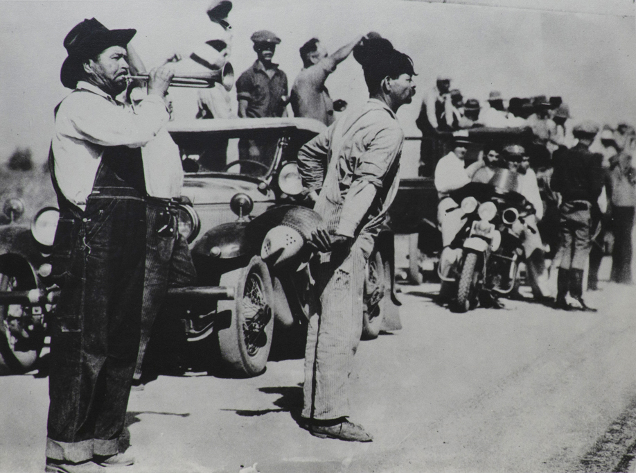 Black and white photograph of of figures standing in front of car which is leading caravan. Man to left in dark hat wearing coveralls is blowing a bugle. Police officer on motor bike in center of picture. More figures standing around in other cars in rear. 