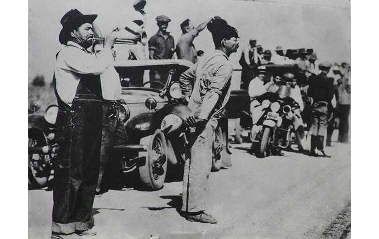 Black and white photograph of of figures standing in front of car which is leading caravan. Man to left in dark hat wearing coveralls is blowing a bugle. Police officer on motor bike in center of picture. More figures standing around in other cars in rear.