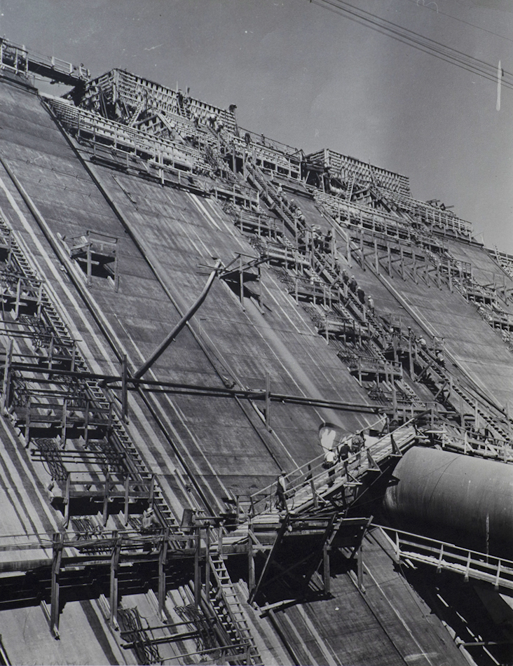 Black and white photograph of construction of the face of a dam. Ladders and working stations. Some hard-hatted workers climbing stairs.