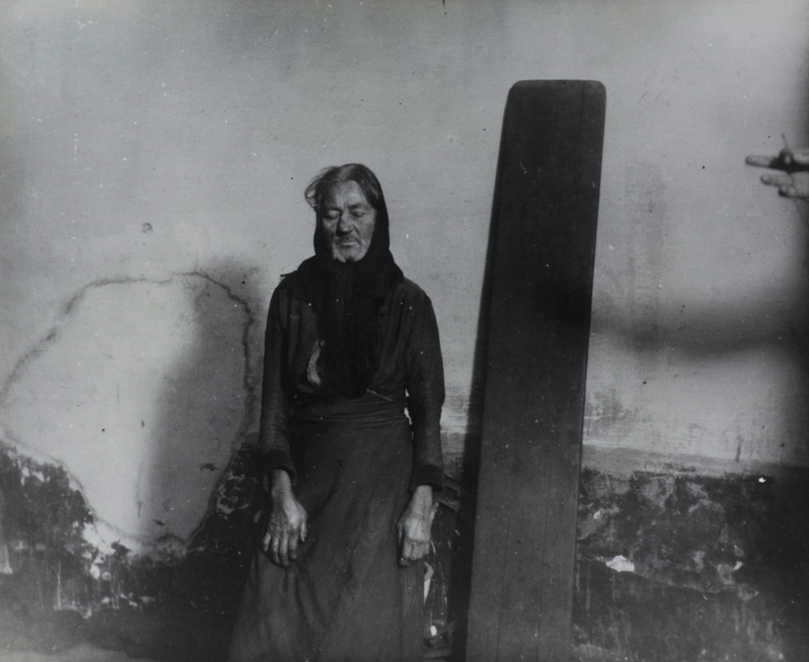 Black and white photograph of figure with eyes closed standing by a plank that leans against the wall. A hand holds a match on the right side of image.