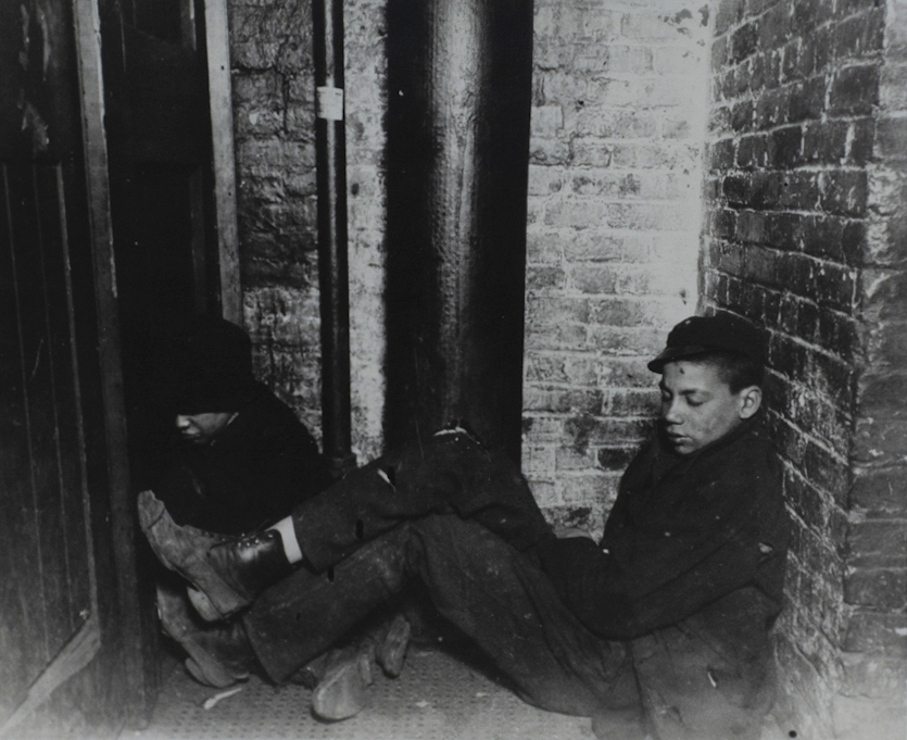Black and white photograph of two young people sleeping in a corner. Brick walls and black pipe in the background.