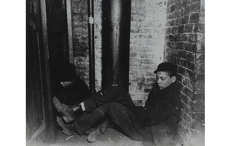 Black and white photograph of two young people sleeping in a corner. Brick walls and black pipe in the background.