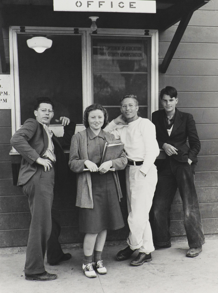 Black and white photograph of a female holding books, three males to her sides. They are standing in front of window labeled 'Office'.