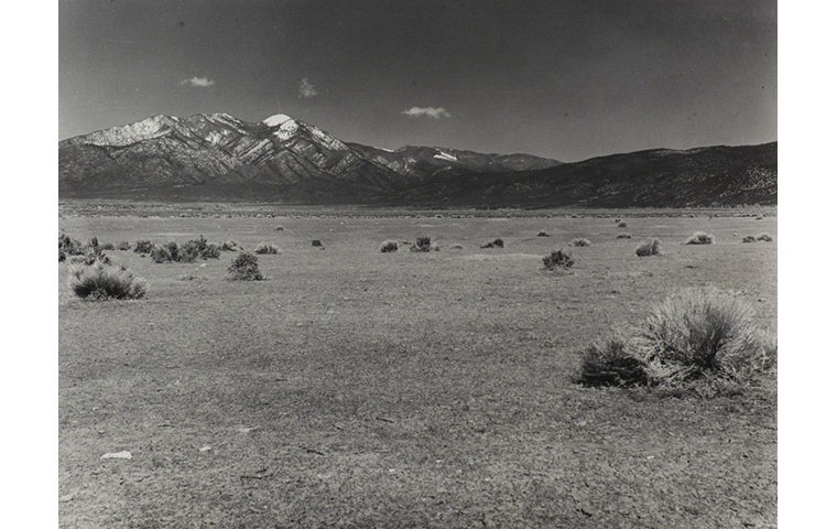 Black and white photograph of an open field land with sagebrush bushes throughout the field. Partially snow covered mountain range in background. Three small puffs of clouds above snow peaks.