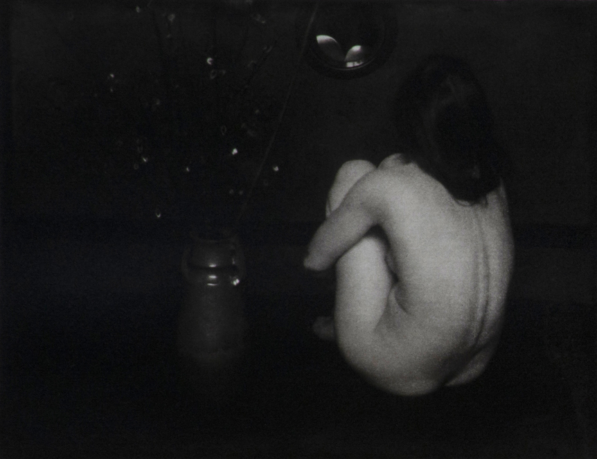 Black and white photograph of nude woman sitting with back toward photographer, clasping her knees. A floral arrangement in a vase to the left. Woman's knees are reflected in a small round mirror in the upper center.