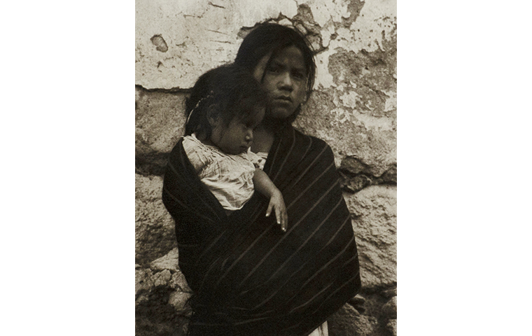 Black and white photograph of young woman in a serape holding a young girl. Stone wall background.