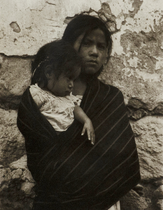 Black and white photograph of young woman in a serape holding a young girl. Stone wall background.