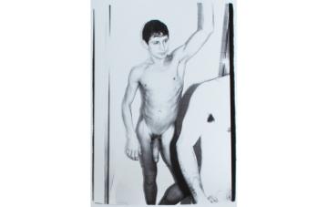 Black and white photograph of male nude wearing a penis ring standing in shower. Left arm raised and looking towards a figure in front of him. Other figure on the right is partially shown from shoulder to pelvis.
