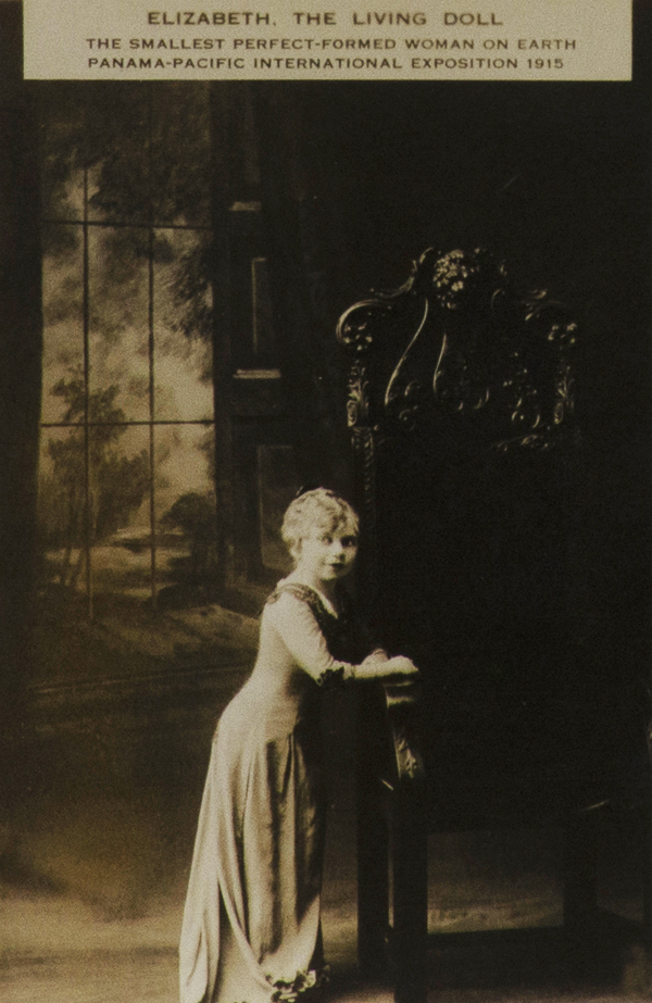 Sepia toned photograph of small petite figure dressed in a long gown standing next to an ornately carved wooden chair.