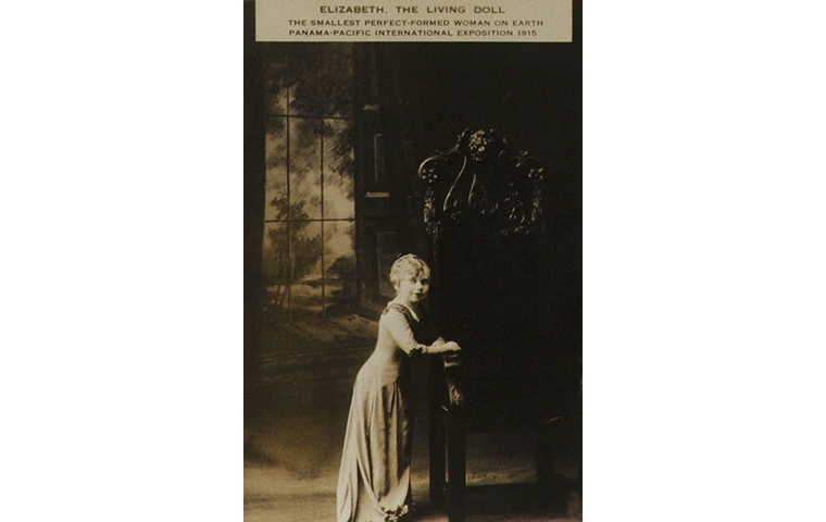 Sepia toned photograph of small petite figure dressed in a long gown standing next to an ornately carved wooden chair.