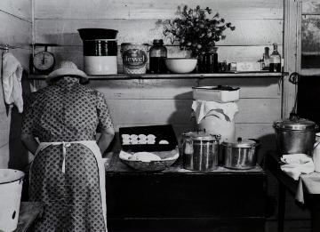 Black and white photograph of back of figure in short sleeve patterned dress with white apron and hat working with dough on a countertop with dough, uncooked biscuits, pots, and pans. 