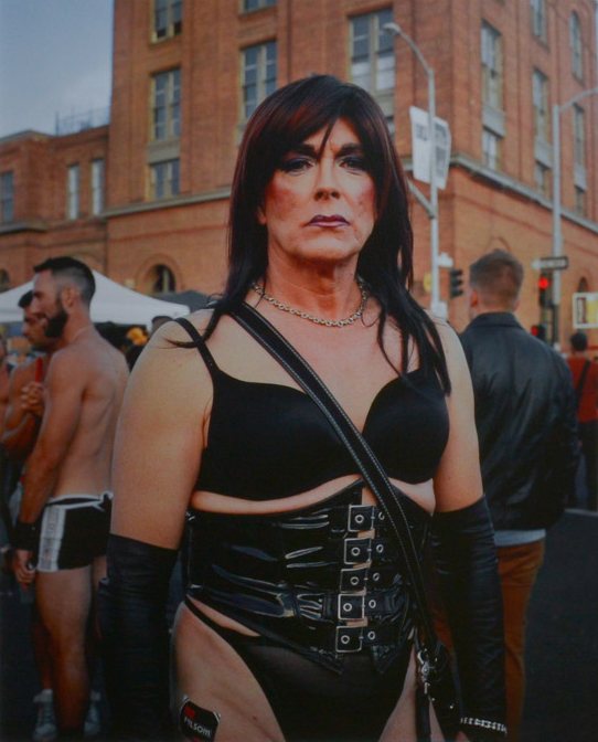 Color photograph of a figure dressed in a black leather bra and corset. Background of people standing at a street festival.