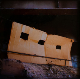Color photograph of a cardboard wall structure with three square cut out for windows. Flat structure is standing with wires attached to underpass structure.