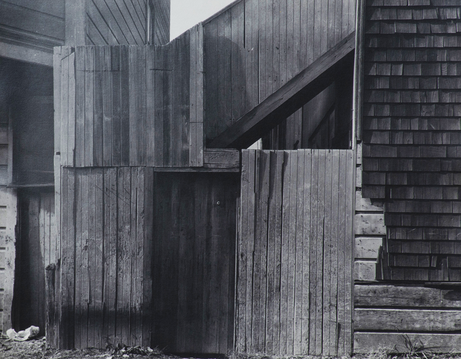 Black and white photograph of the outside of an old building. Wood vertical fence boards in front of a staircase. Shingles on the right side are partially gone. The wooden gate door is shaded.