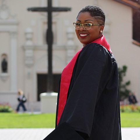 Graduate poses for a photo in front of the Mission Church in regalia 