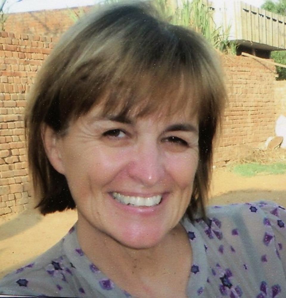 A person smiling outdoors with trees and a brick wall background.