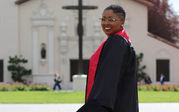 Person in graduation attire on a college campus, smiling at the camera. image link to story