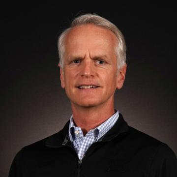 A person in a jacket and collared shirt with a dark background.