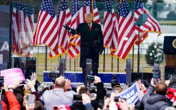 A person speaking on stage with American flags in the background.