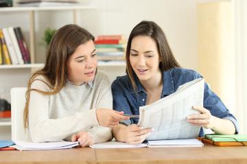 Two female students reading a newspaper together at a table.
