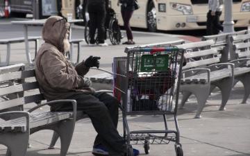 Man sitting on public bench with shopping cart filled with personal belongings.