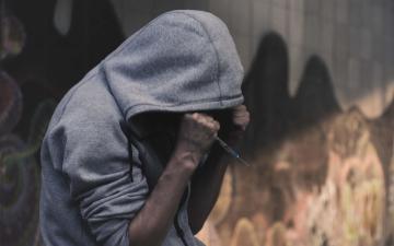 A person wearing a hoodie is sitting outdoors against a graffiti filled concrete wall and is holding their head with fisted hands. One hand holds a hypodermic needle.