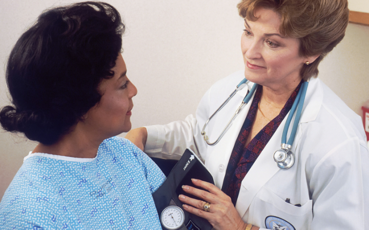 A woman with a stethoscope and wearing a medical coat talking to another woman wearing a patient's gown.