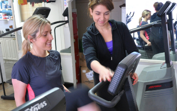 A woman in a gym setting sitting on a recumbent cycling fitness machine and another woman explaining how the machine monitor functions.