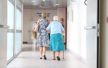 A woman assists an elderly woman walking in a hospital corridor.