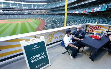 People receiving vaccines at a baseball stadium with a sign reading COVID Vaccine This Way. image link to story