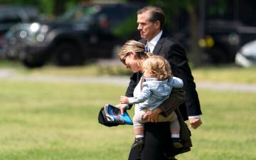 Hunter Biden carrying a young child while walking outdoors in a grassy area. image link to story