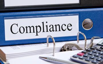 Close-up of a binder labeled Compliance on a desk with a calculator. image link to story