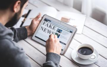 Person looking at tablet screen displaying Fake News headline with coffee on table. image link to story