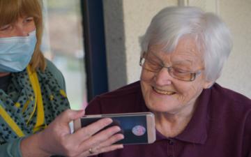 A senior person looking at a smartphone, assisted by a person wearing a mask. image link to story