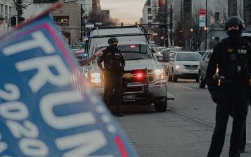 People with a Trump 2020 banner and police van in urban area.
