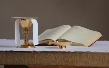 Open Bible with glass of wine and bread on a table. image link to story