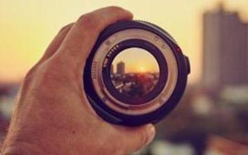 A person holds a camera lens to view the setting sun. 