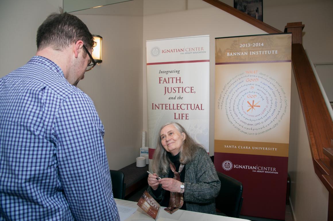Alt text: Author signing a book for a person at a table, banner in the background.