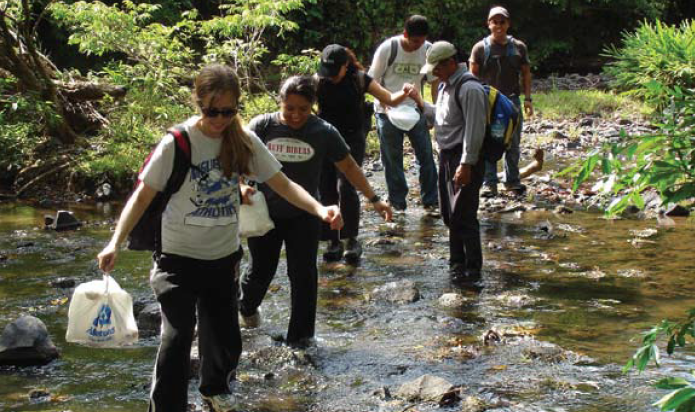 Group of people walking across a shallow stream carrying bags and equipment.